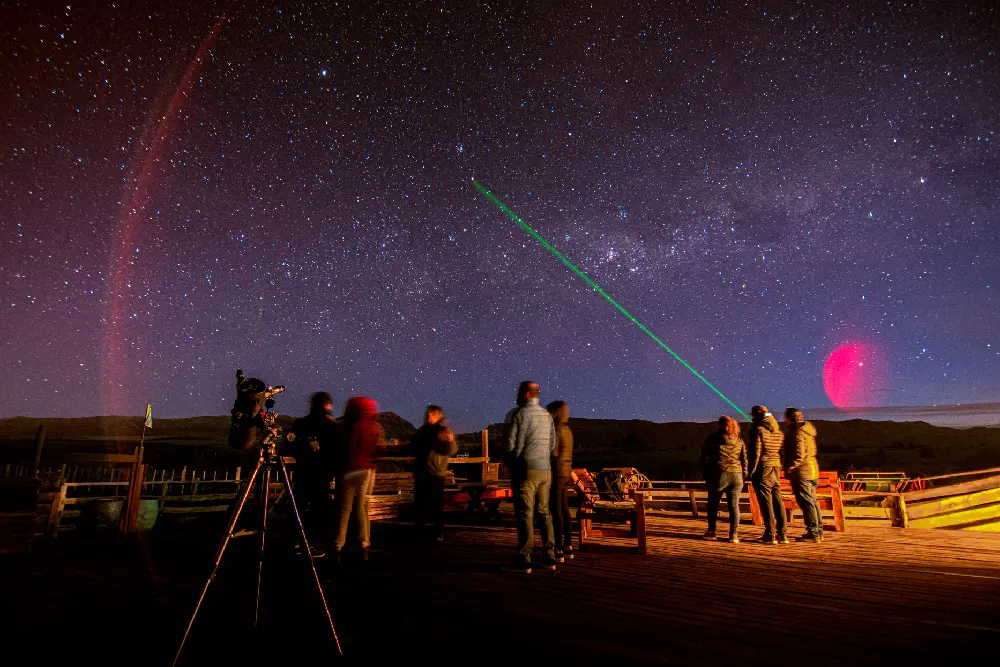Un grupo de personas abrigadas en un deck de madera mira el cielo estrellado mientras un guía señala con un láser verde, junto a un telescopio