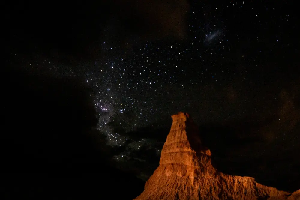 Un pico de roca rojiza iluminado de noche, bajo un cielo oscuro con muchas estrellas