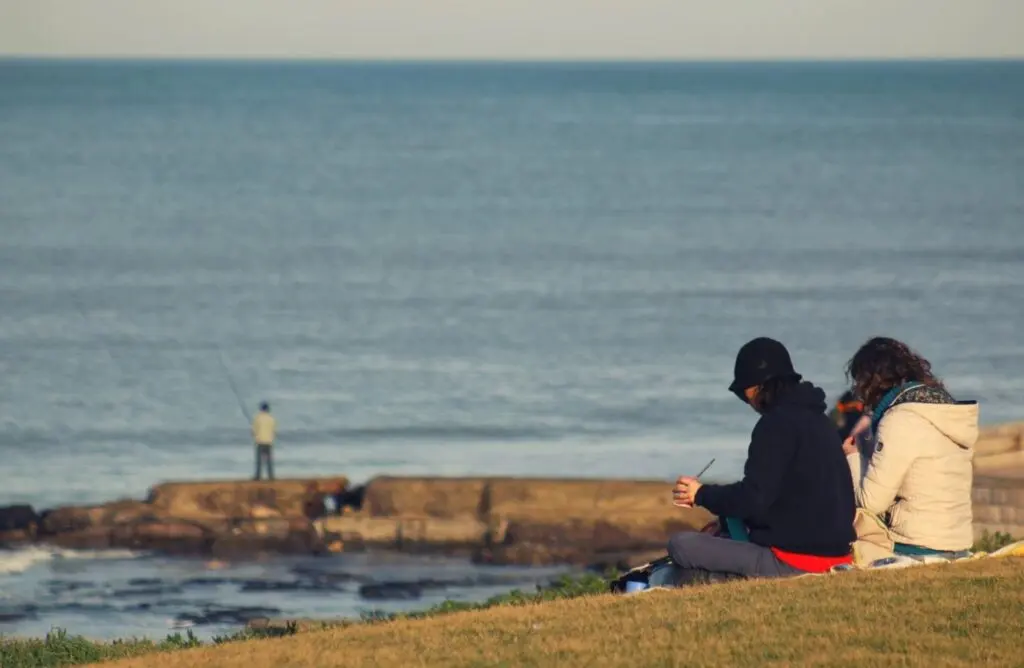Dos personas sentadas en el pasto frente al mar, con un pescador parado sobre las rocas de la costa