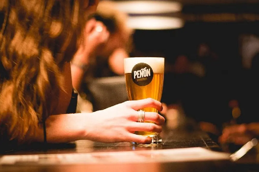 Una mujer apoyada en la barra de un bar sostiene un vaso de cerveza rubia