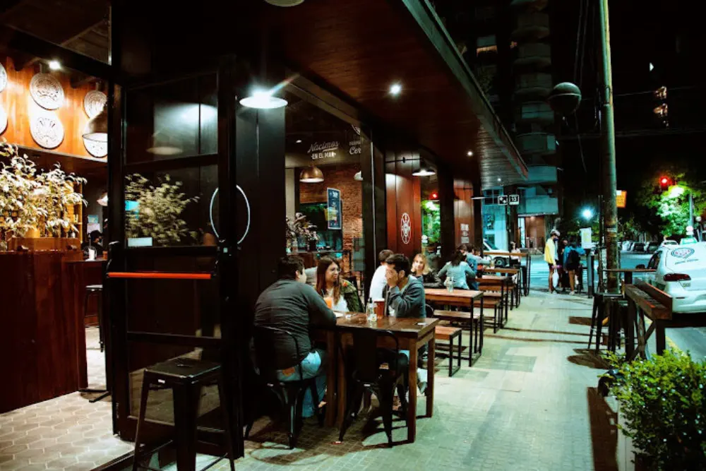 Un bar en una esquina de noche, con grupos de amigos sentados en mesas de madera sobre la vereda