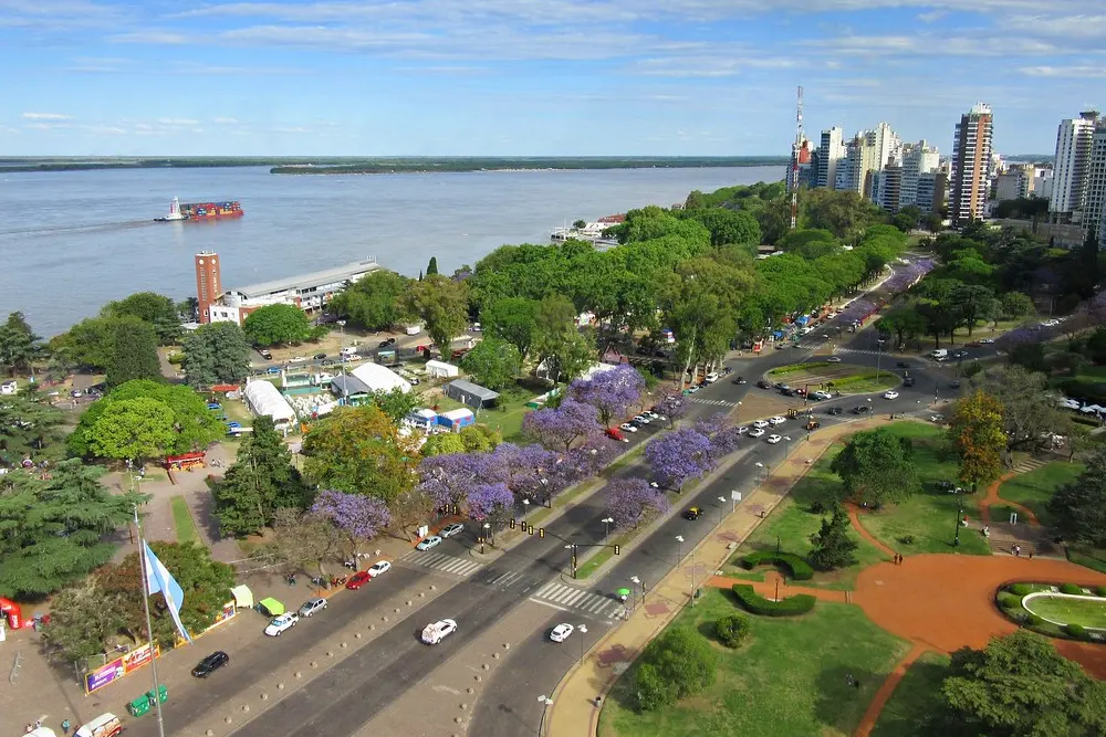 Vista aérea de la costanera central de Rosario, con jacarandás en flor sobre la avenida, un parque y un buque de carga en el río