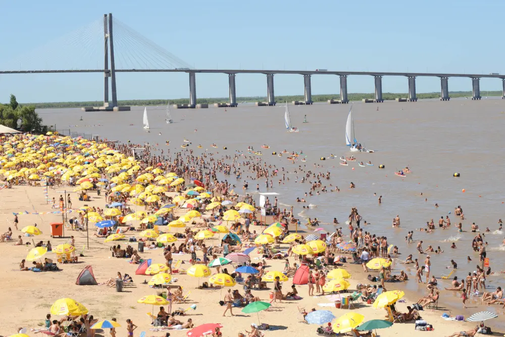 Una playa de río llena de sombrillas amarillas y bañistas en el agua, con veleros y el puente Rosario-Victoria al fondo