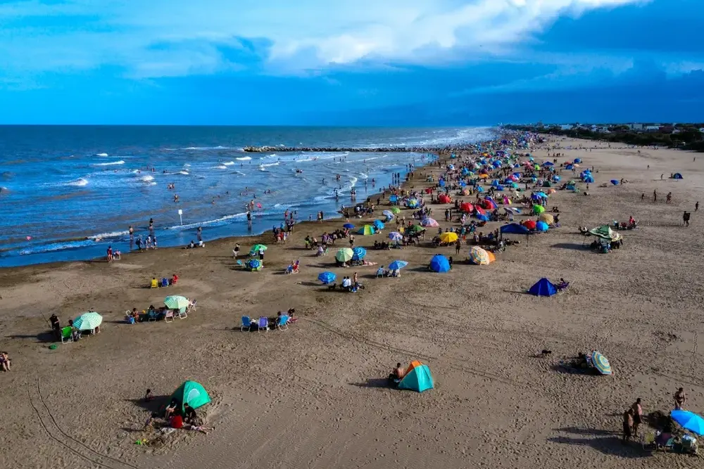 Vista aérea de la playa con sombrillas y carpas de colores, gente en el agua y una escollera al fondo