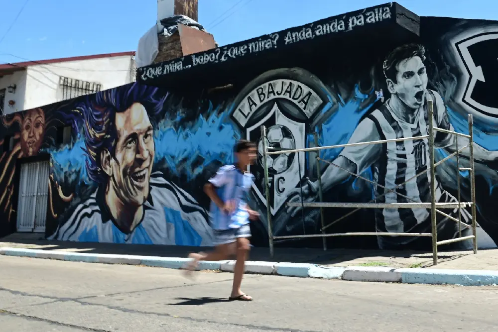 Un chico con la camiseta argentina pasa corriendo frente a un mural de barrio con dos retratos de Messi y el escudo de un club