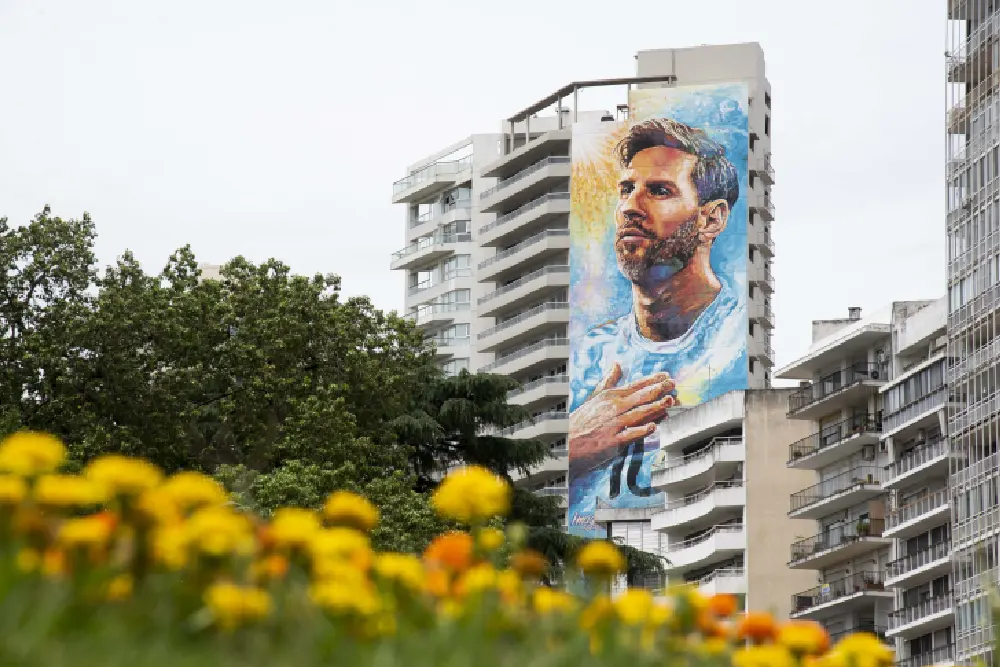 Mural de Messi con la mano en el pecho en la medianera de un edificio, visto desde un parque con flores amarillas