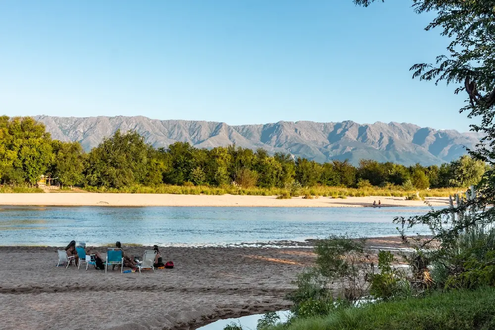 Un grupo sentado en reposeras sobre una playa de arena junto a un río ancho, con una arboleda y las sierras detrás
