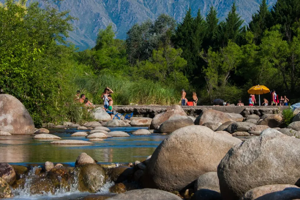 Río de poca profundidad entre grandes piedras redondeadas, con bañistas y una sombrilla amarilla en la orilla