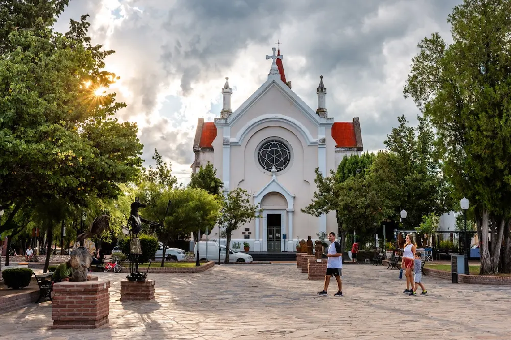 Plaza empedrada con esculturas de hierro y una iglesia blanca de techos rojos al fondo, bajo un cielo con nubes