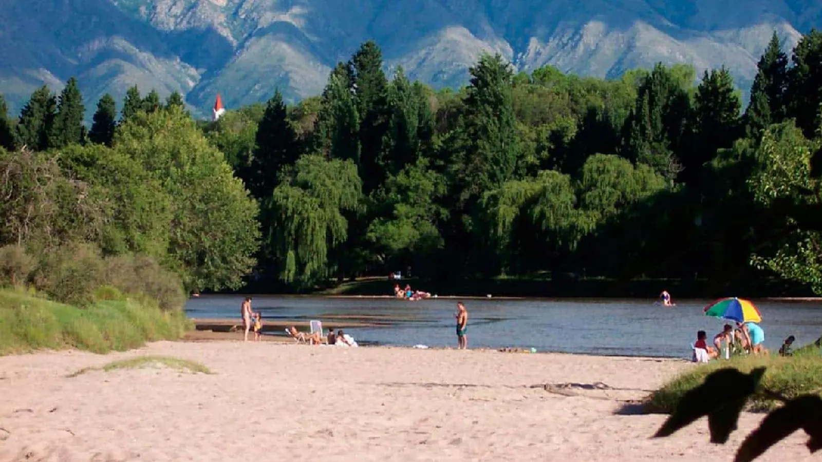 Playa de arena junto a un río con gente bañándose, una arboleda tupida, la torre roja de una iglesia y las sierras de fondo