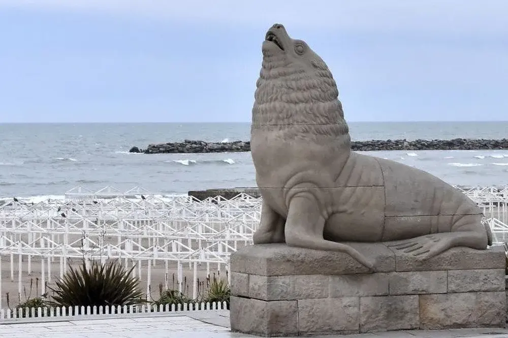 Escultura de piedra de un lobo marino en la rambla de Mar del Plata, con las carpas de un balneario y el mar detrás