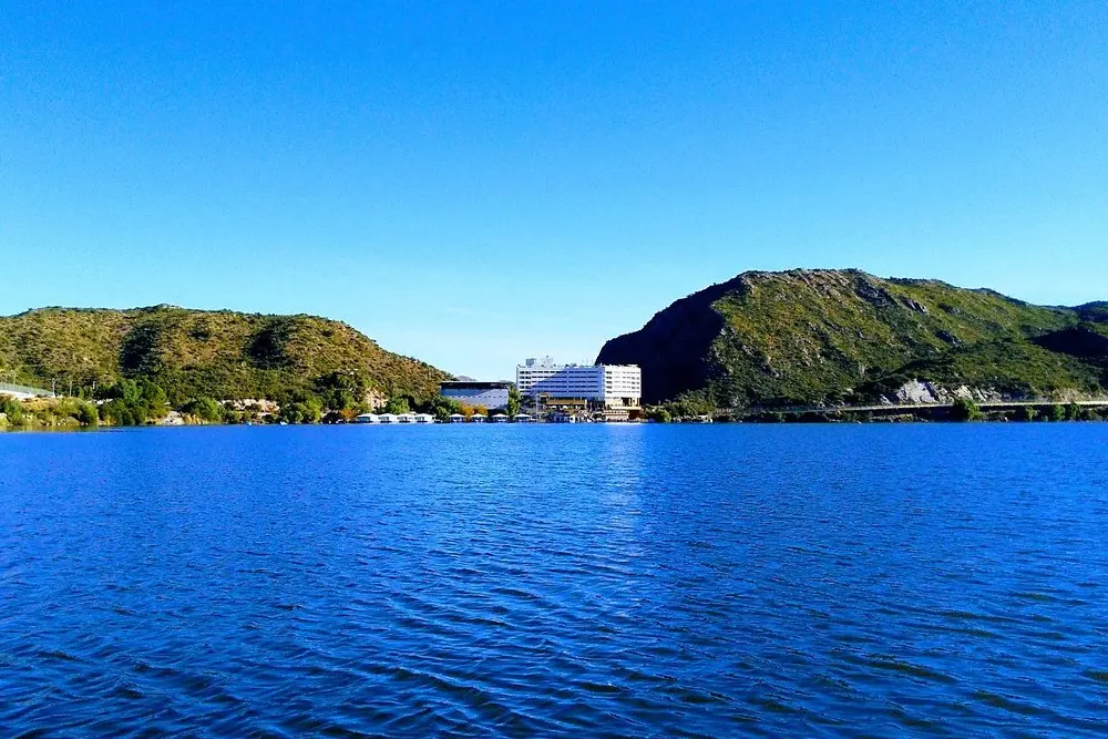 Lago de agua azul entre dos cerros verdes, con un edificio blanco de varios pisos en la orilla