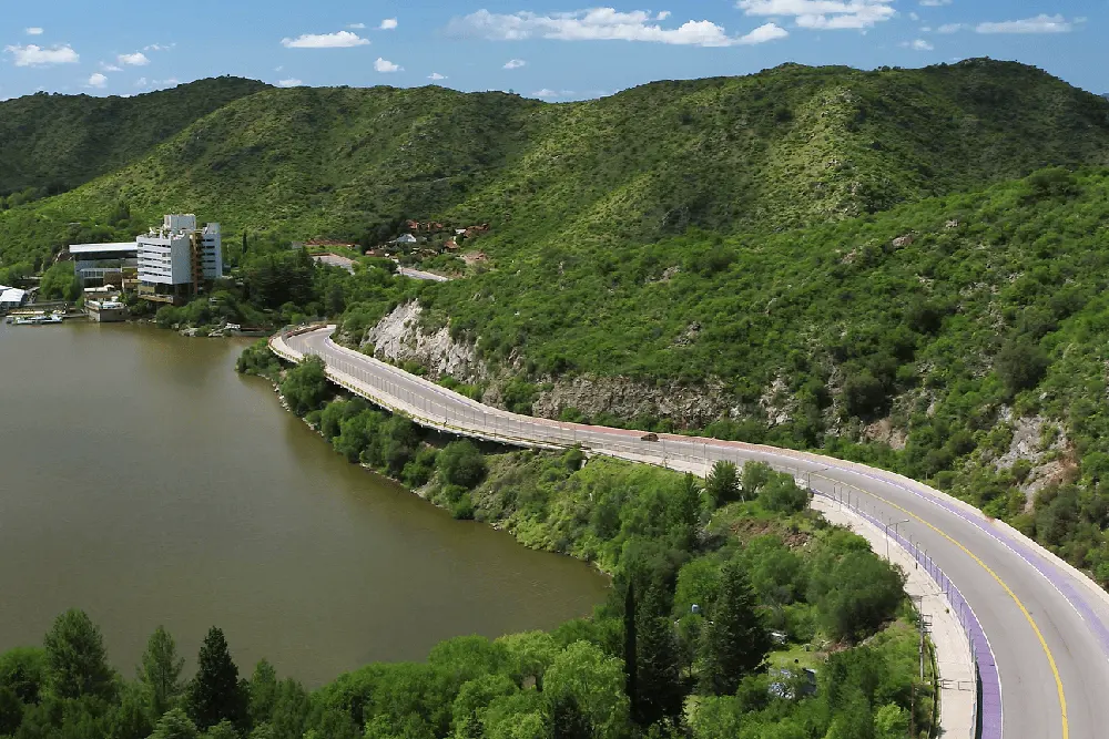 Ruta asfaltada que bordea un lago entre cerros cubiertos de vegetación, con un hotel blanco en la orilla