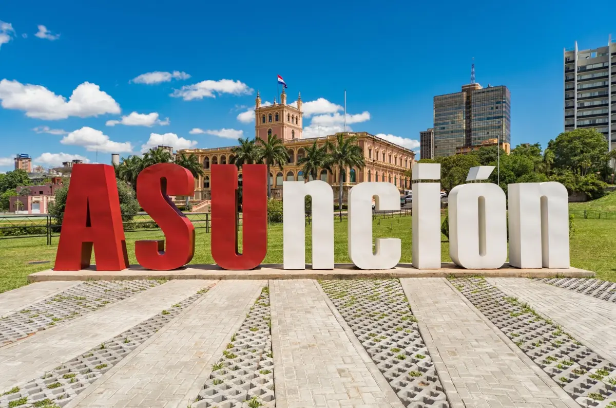 Letras gigantes rojas y blancas que forman la palabra Asunción, con el Palacio de López y palmeras detrás