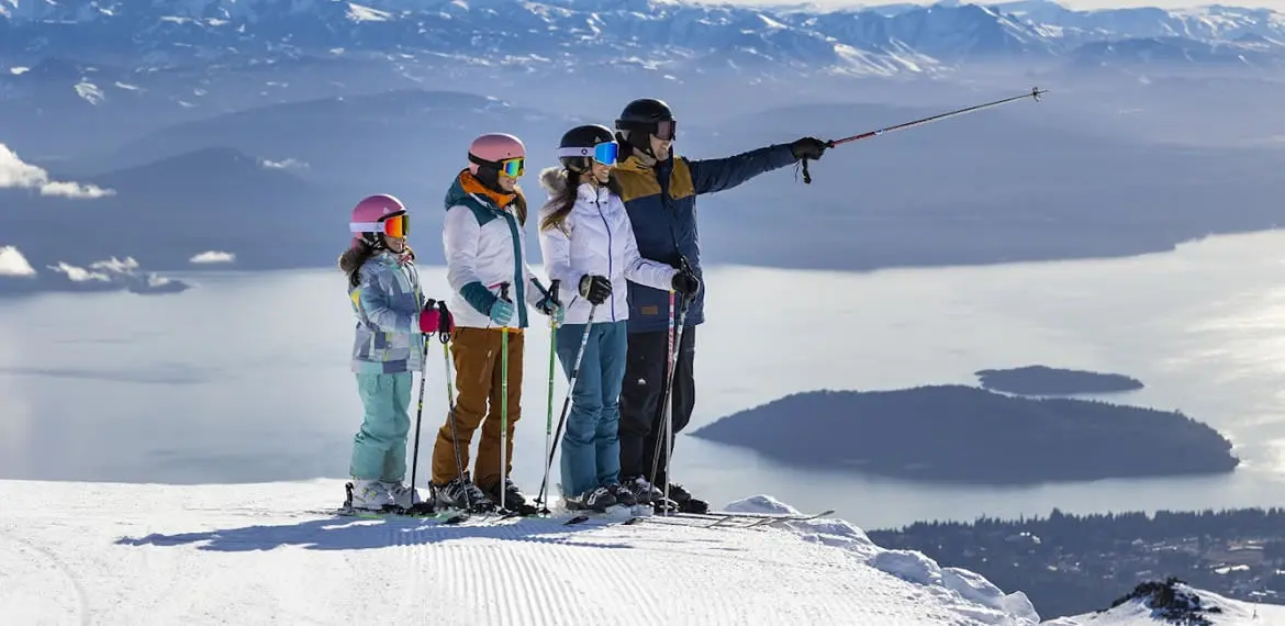Una familia con equipo de esquí en lo alto de una pista nevada, con el lago y sus islas abajo
