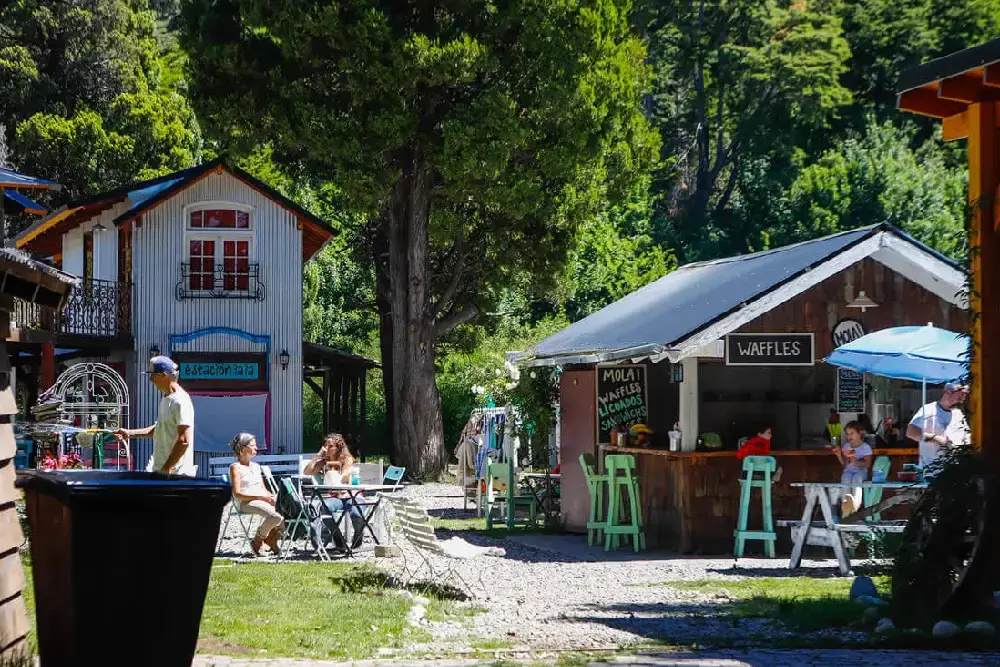 Casas de madera y chapa, un puesto de waffles y mesas al sol entre árboles altos
