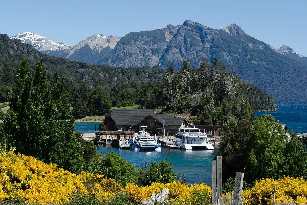 Muelle con dos catamaranes en una bahía de agua turquesa, rodeada de bosque y cerros con nieve