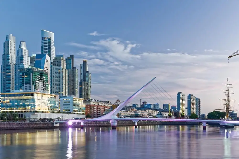 El Puente de la Mujer iluminado sobre el dique de Puerto Madero al atardecer, con las torres vidriadas y un velero de fondo