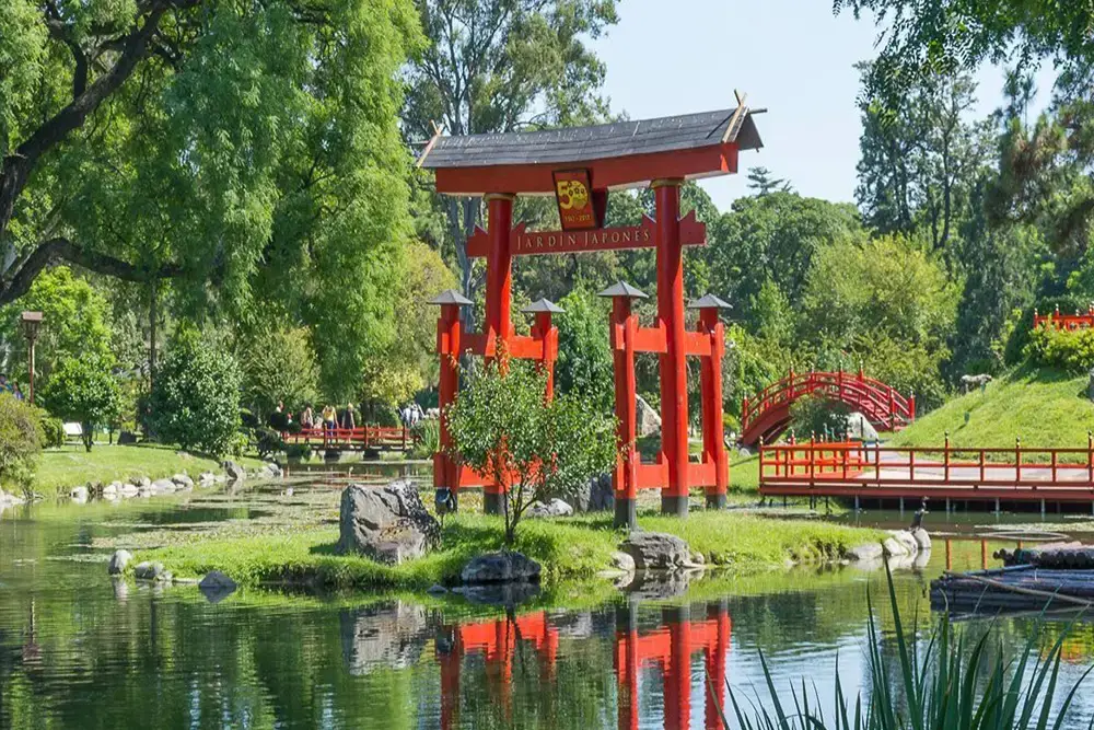 Portal rojo de estilo japonés junto a un estanque, con un puente rojo curvo y árboles frondosos detrás