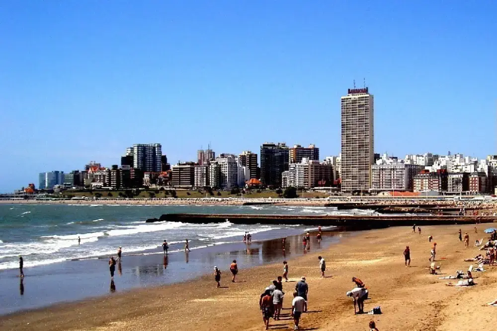 Playa de arena con gente caminando por la orilla y los edificios de Mar del Plata al fondo, bajo un cielo despejado