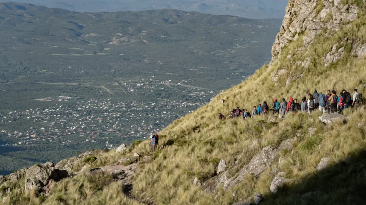 Un grupo de caminantes en fila por la ladera de pastizales del Cerro Uritorco, con el pueblo y el valle abajo