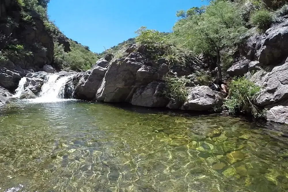 Una pequeña cascada cae entre rocas sobre un piletón de agua transparente en un río serrano
