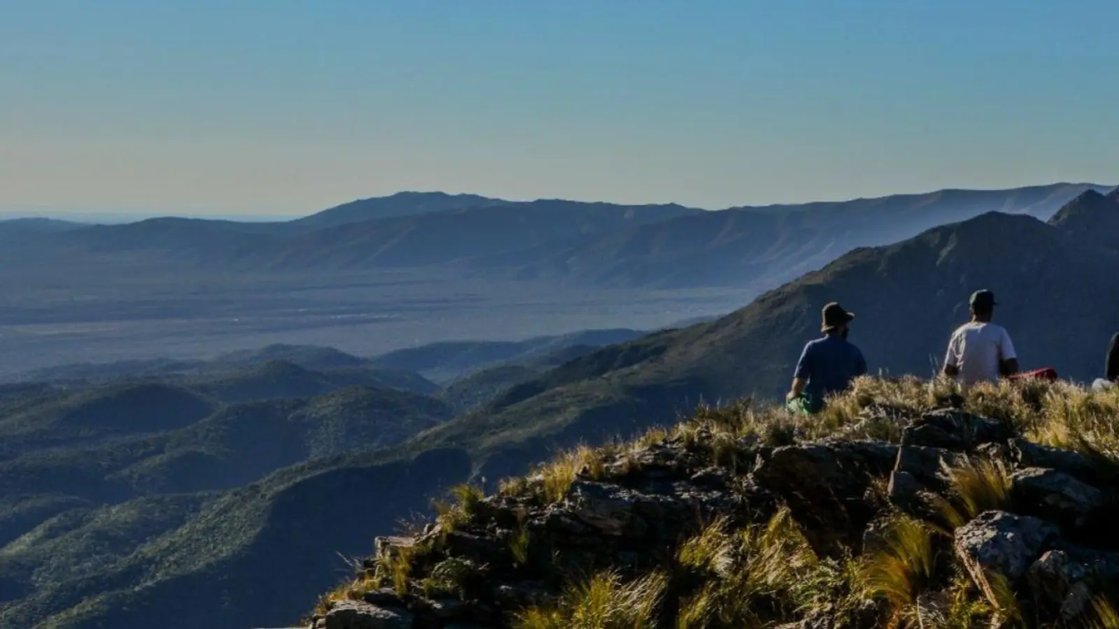 Caminantes sentados en una cumbre serrana de Capilla del Monte, mirando el valle y los cordones de sierras al atardecer