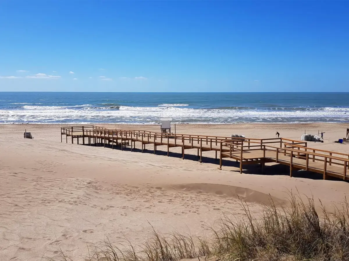 Pasarela de madera que baja entre médanos con pastizales hasta una playa ancha, con un puesto de guardavidas y el mar de fondo