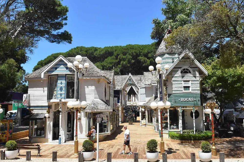 Galería comercial de casitas de madera blancas y celestes de estilo victoriano, con faroles y pinos detrás
