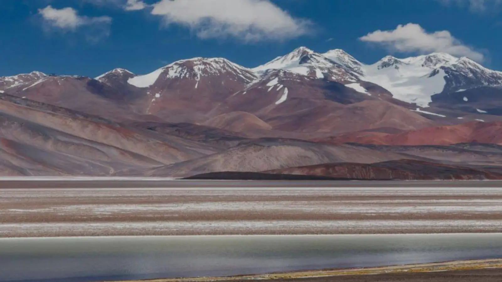 Salar de la Puna catamarqueña con una laguna en primer plano y cerros nevados de tonos rojizos al fondo