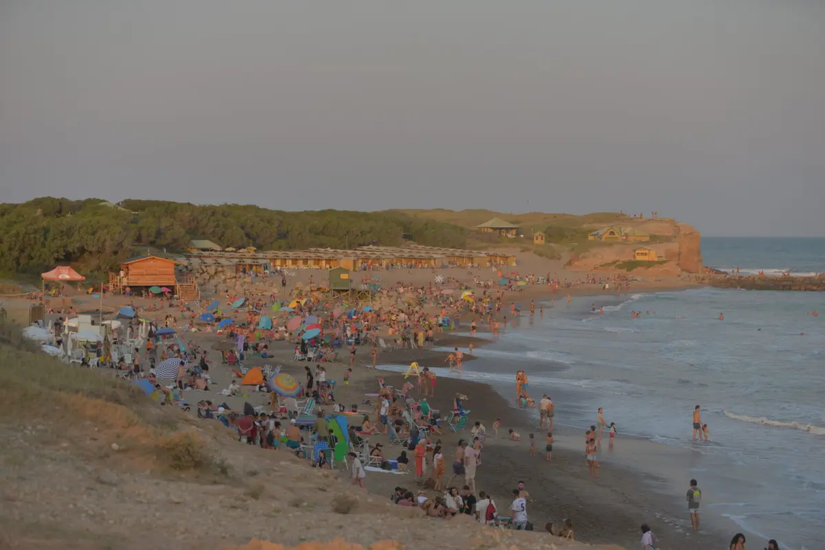 Playa con mucha gente al atardecer, con paradores de madera, un acantilado al fondo y el mar con olas suaves
