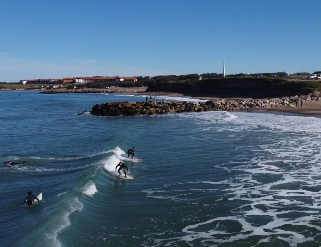 Surfistas con traje de neoprene montando una ola junto a una escollera de piedras, con los edificios de techos rojos de la Unidad Turística en la costa