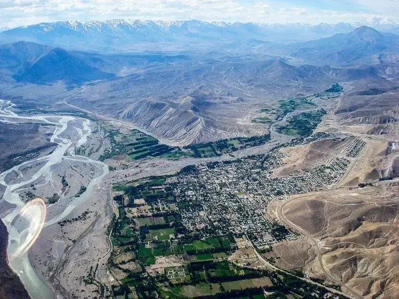 Vista aérea de una ciudad en un valle verde junto a un río ancho, rodeada de cerros áridos y cordillera nevada al fondo