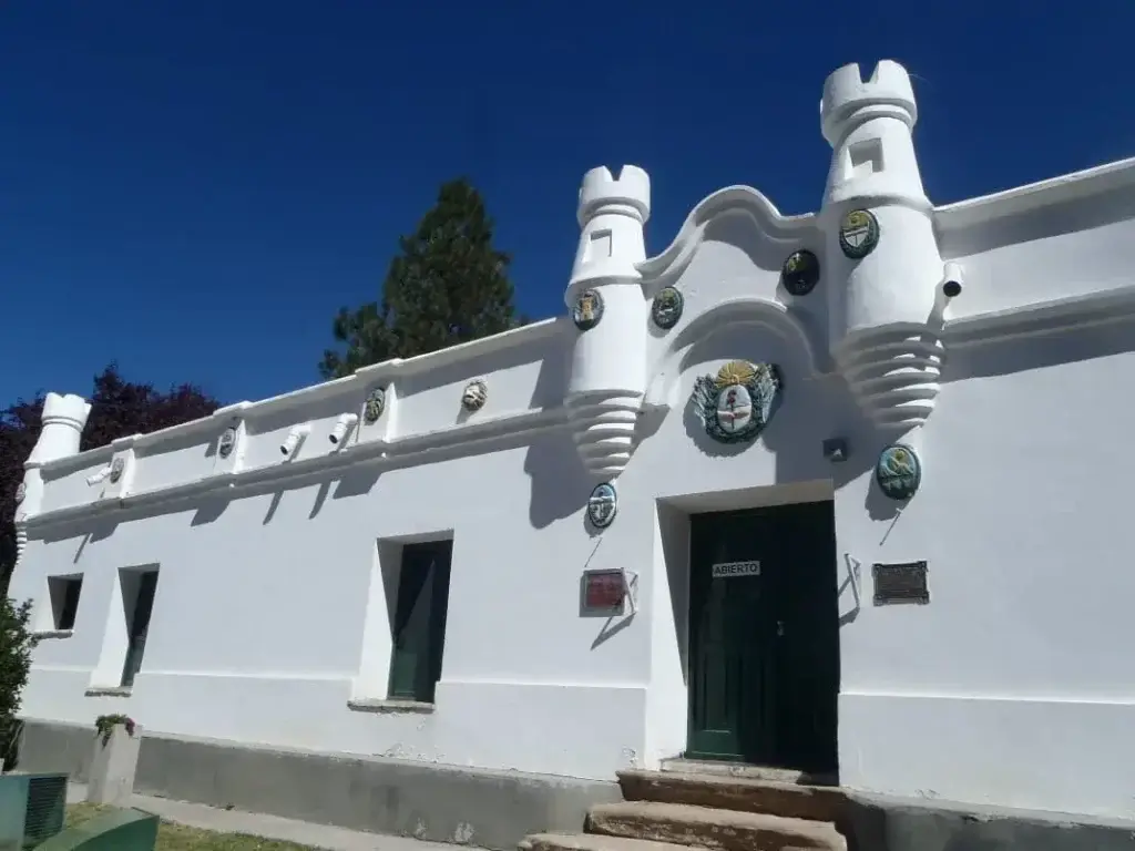 Fachada blanca de un edificio histórico con dos torrecillas almenadas, escudos de colores y una puerta verde