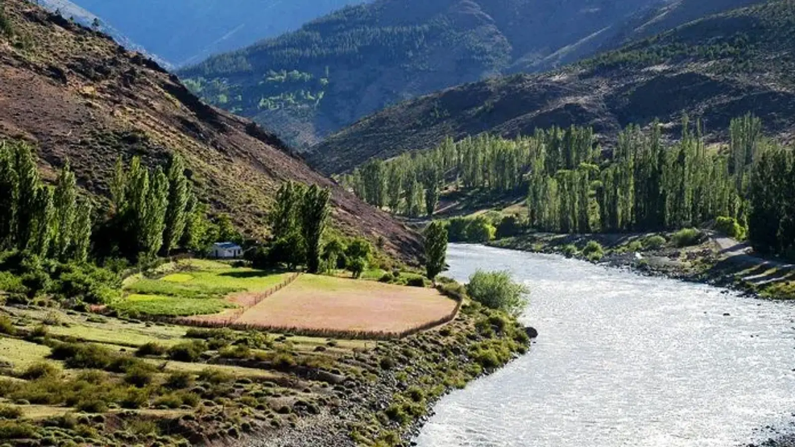 Río de aguas claras que corre por un valle de montaña, con chacras y álamos en la orilla