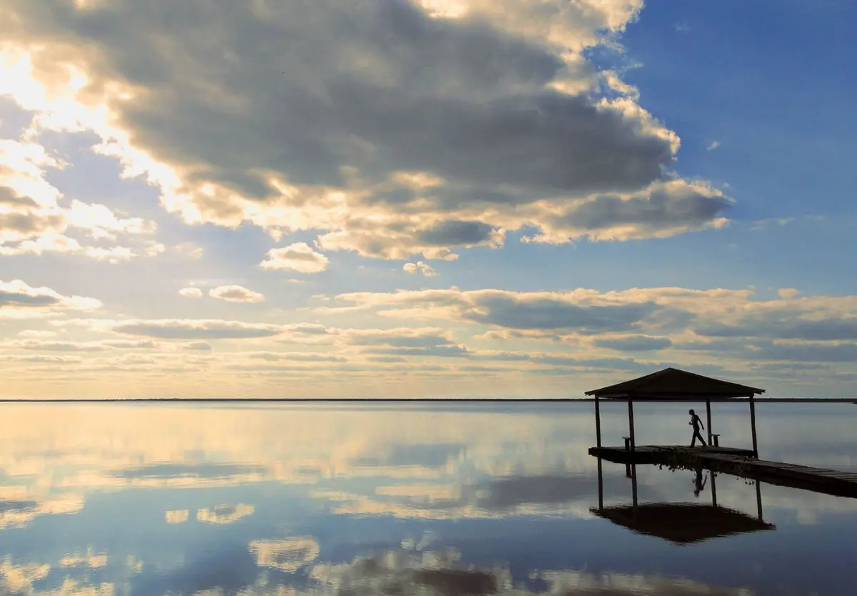 Un muelle de madera con techo sobre una laguna enorme y quieta que refleja las nubes del atardecer, con una persona caminando por el muelle