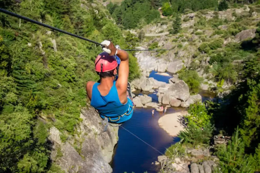 Una persona con casco cruza en tirolesa sobre un río que corre entre rocas y un bosque de pinos
