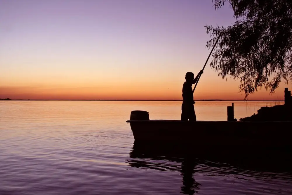 Silueta de un lanchero que empuja una lancha con una vara en una laguna al atardecer, con el cielo violeta y naranja
