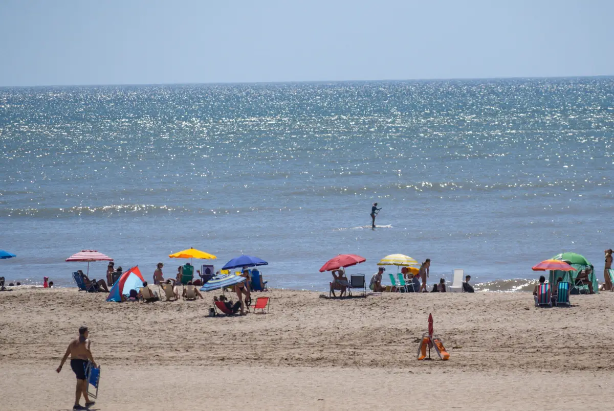 Playa con sombrillas de colores y gente en reposeras, un hombre parado sobre una tabla de remo en el mar y el agua brillando al sol