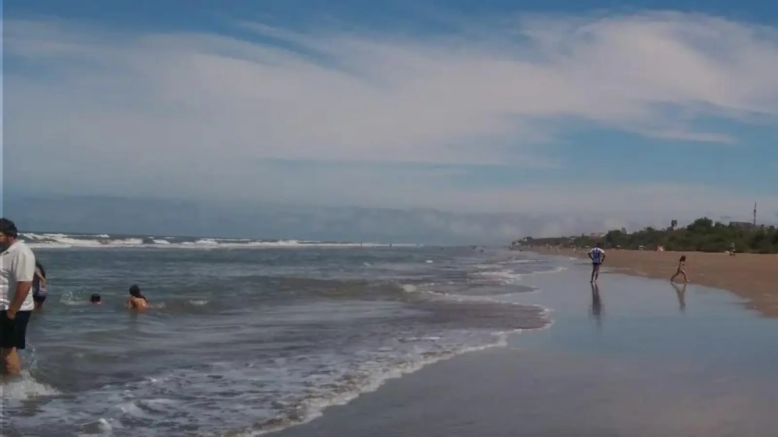 Playa ancha de Costa Chica con gente bañándose en la orilla, el cielo con nubes altas y una franja de árboles sobre los médanos