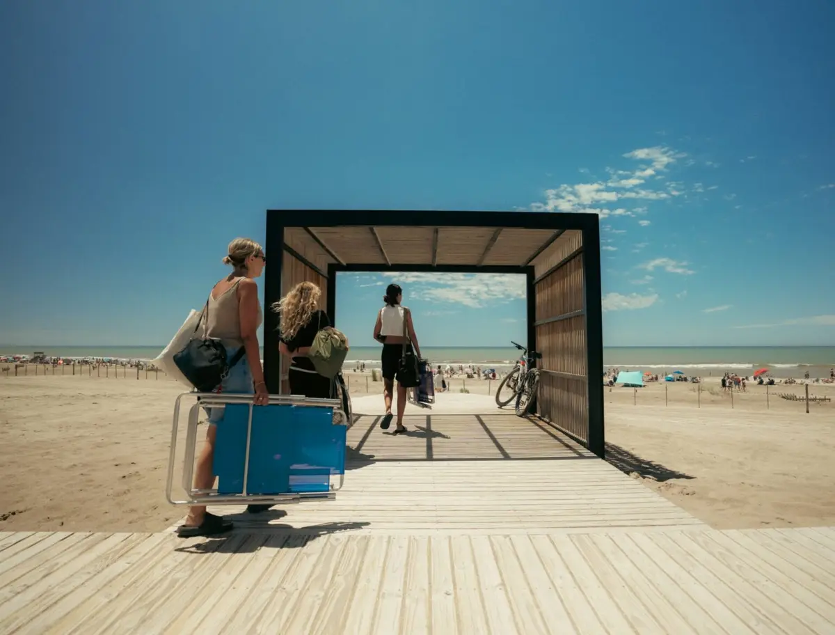 Tres chicas con reposeras y bolsos entran a la playa por una pasarela de madera con pérgola, con el mar y las sombrillas al fondo