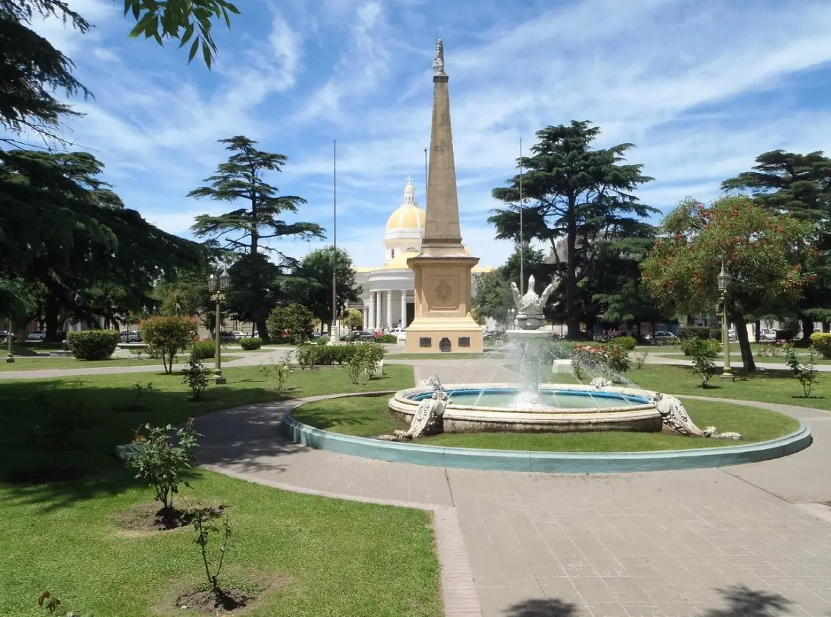 Plaza con una fuente y una pirámide alta coronada por una estatua, y una iglesia de cúpula amarilla al fondo
