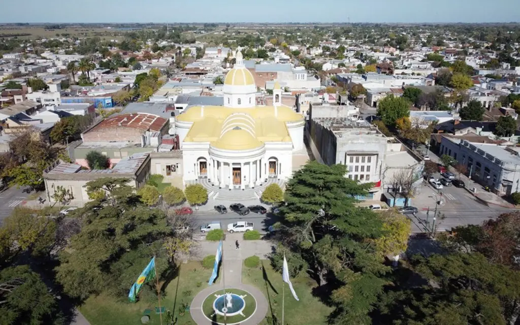 Vista aérea de una iglesia blanca de cúpulas amarillas frente a una plaza arbolada, rodeada por las casas bajas de la ciudad