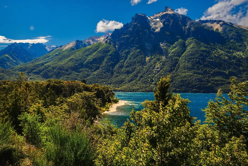 Lago de agua azul con una playa angosta, rodeado de bosque verde y de montañas escarpadas bajo el cielo despejado