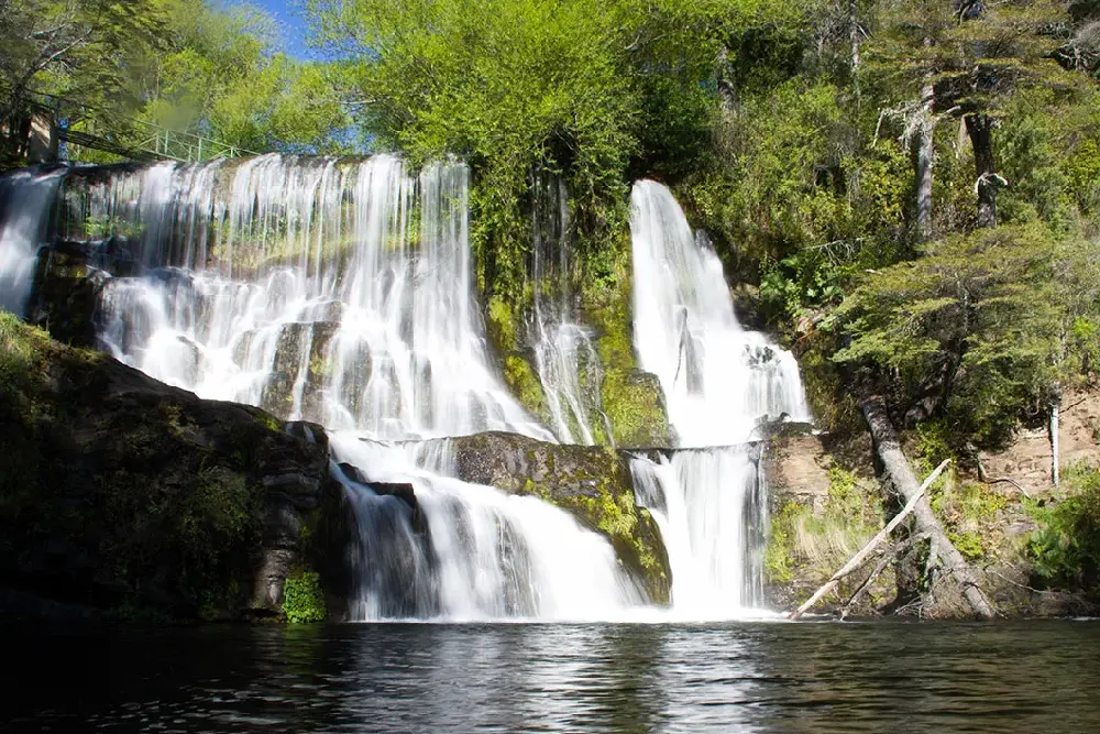 Cascada de varios saltos que cae entre rocas cubiertas de musgo y vegetación hacia una laguna oscura