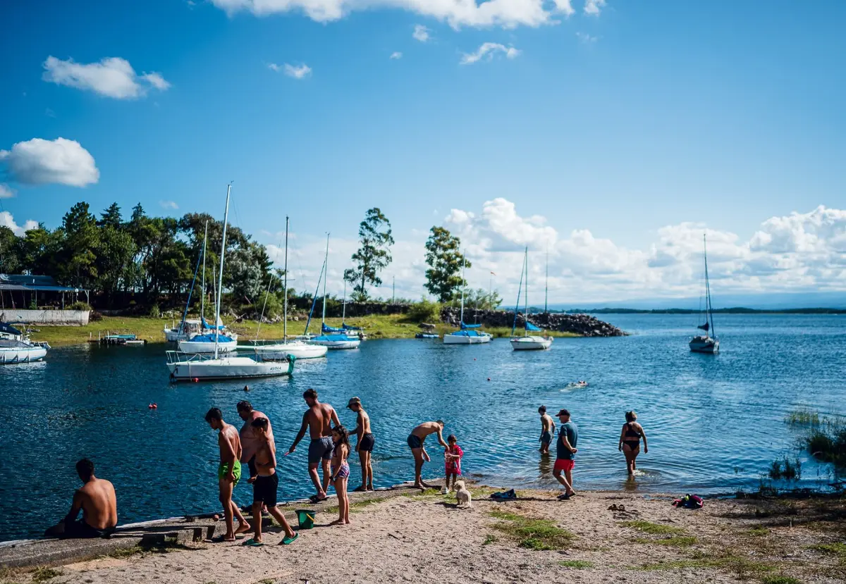 Familias bañándose en la orilla de un lago azul, con veleros amarrados, árboles y sierras bajas a lo lejos