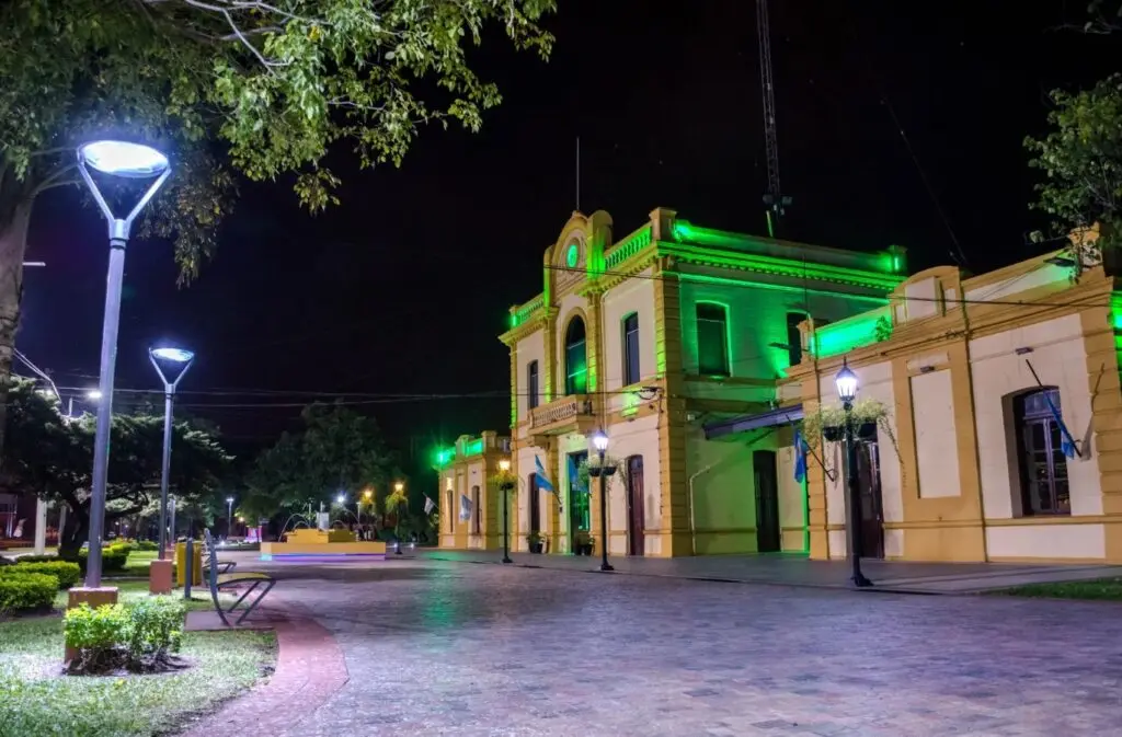Edificio histórico de fachada amarilla iluminado de noche con luces verdes, frente a un paseo peatonal con faroles