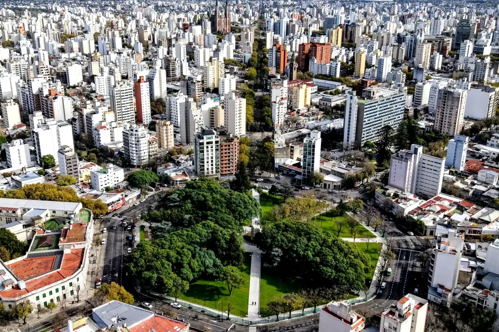 Vista aérea de una plaza arbolada del centro de La Plata rodeada de edificios, con las torres de la Catedral al fondo