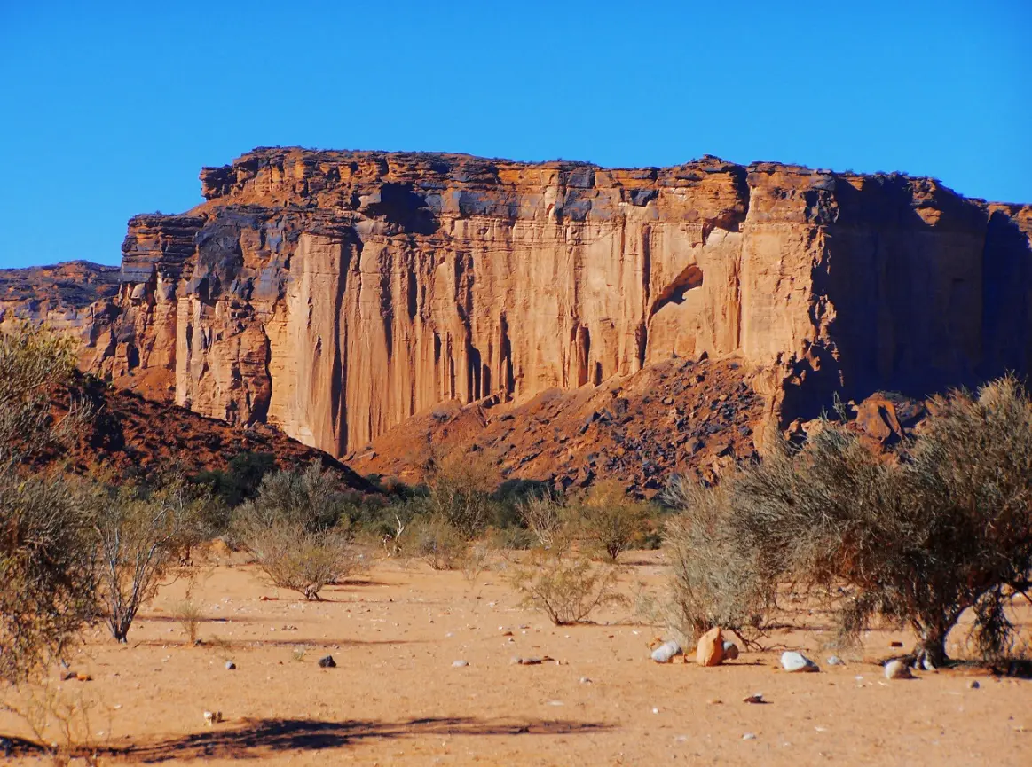 Un gran paredón de roca rojiza con grietas verticales se levanta sobre un suelo de arena con arbustos secos, bajo un cielo azul