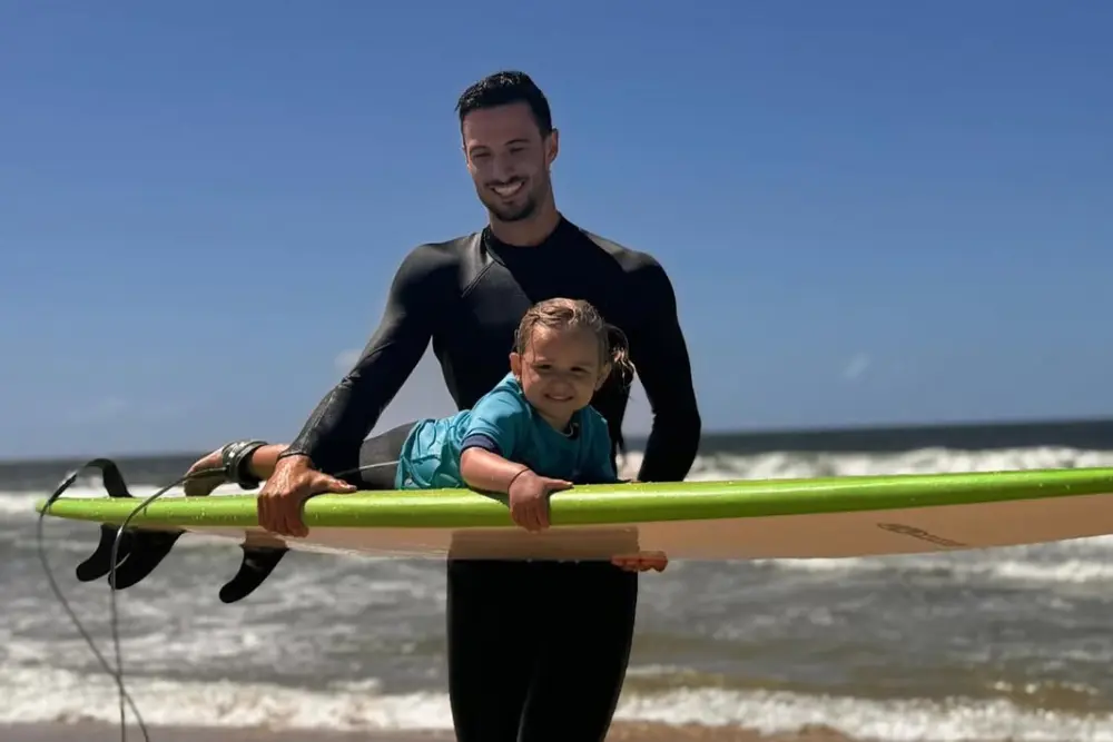 Instructor de surf con traje de neoprene sosteniendo una tabla verde con una nena arriba, en la orilla del mar
